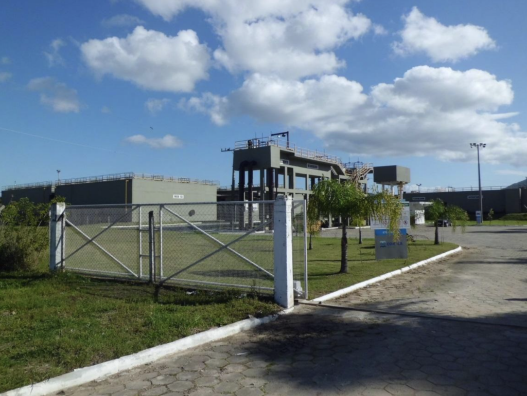 Entrada da estação de tratamento de água com portão e tanques ao fundo, sob céu nublado.